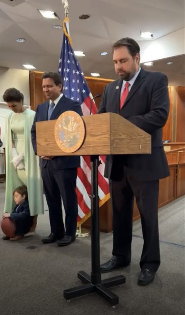 Erik Dellenback praying at a podium bearing the seal of Florida, with Governor and First Lady DeSantis behind him