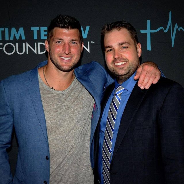 Tim Tebow with his arm around Erik Dellenback in front of a Tim Tebow Foundation backdrop